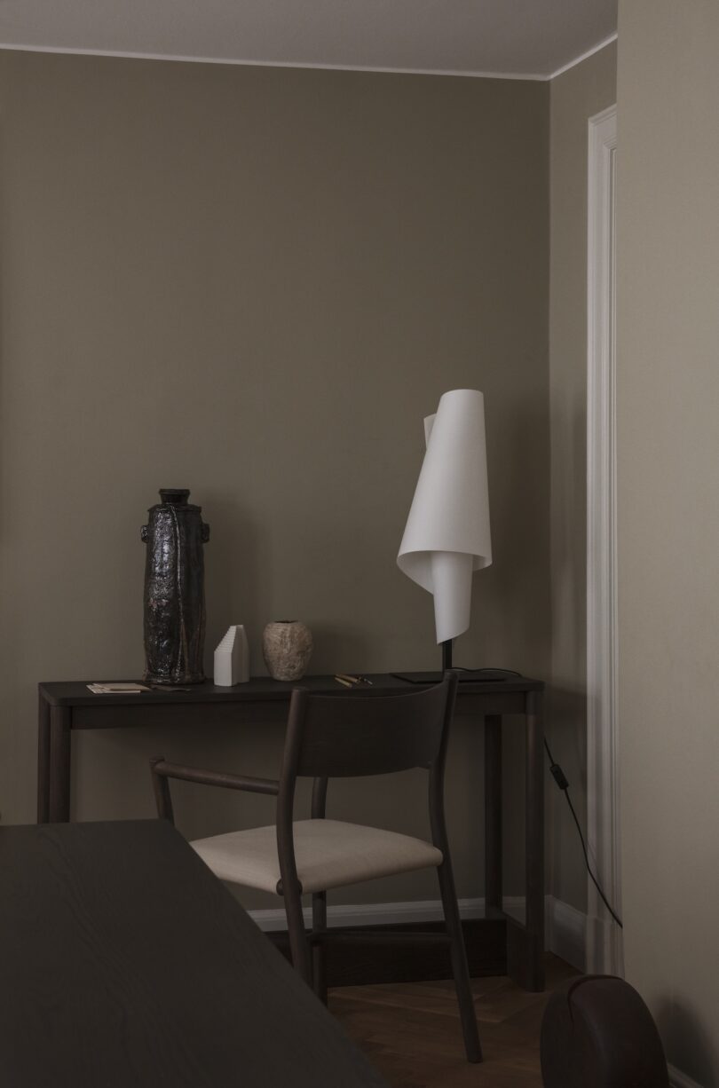 A minimalist desk setup with a wooden chair, ceramic vase, small sculptures, and a modern white table lamp against taupe walls.