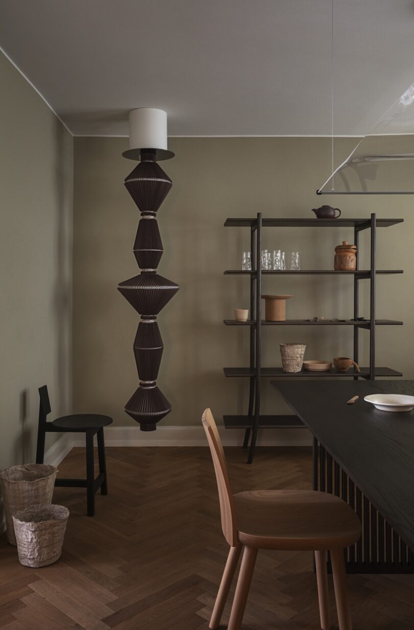 Minimalist dining room with wooden furniture, open shelving holding pottery and glassware, herringbone floor, two baskets, and a sculptural hanging light fixture.