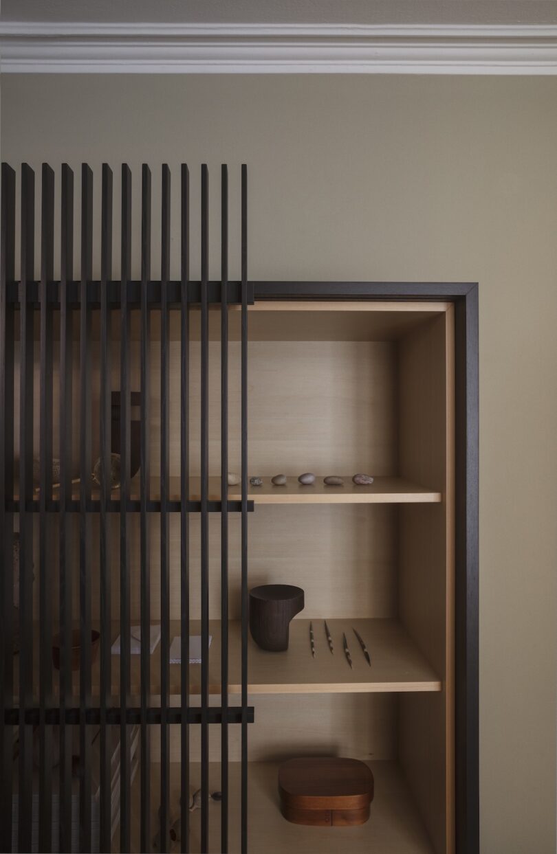 A minimalist wooden shelf with scattered small objects, partially covered by vertical black slats, against a beige wall with white crown molding.
