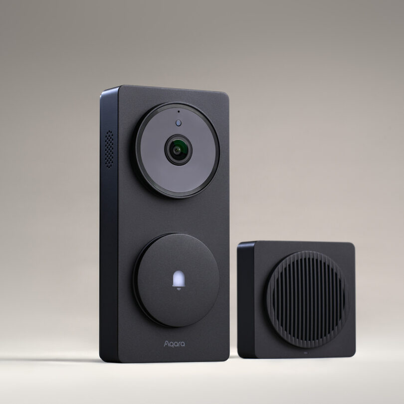 A black smart doorbell camera with a circular button and a matching square speaker unit are displayed against a plain background.