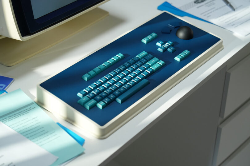 Teclado de computadora MDR Dasher de estilo retro con teclas azules y turquesas, una gran bola de seguimiento negra y documentos dispersos en una mesa blanca.