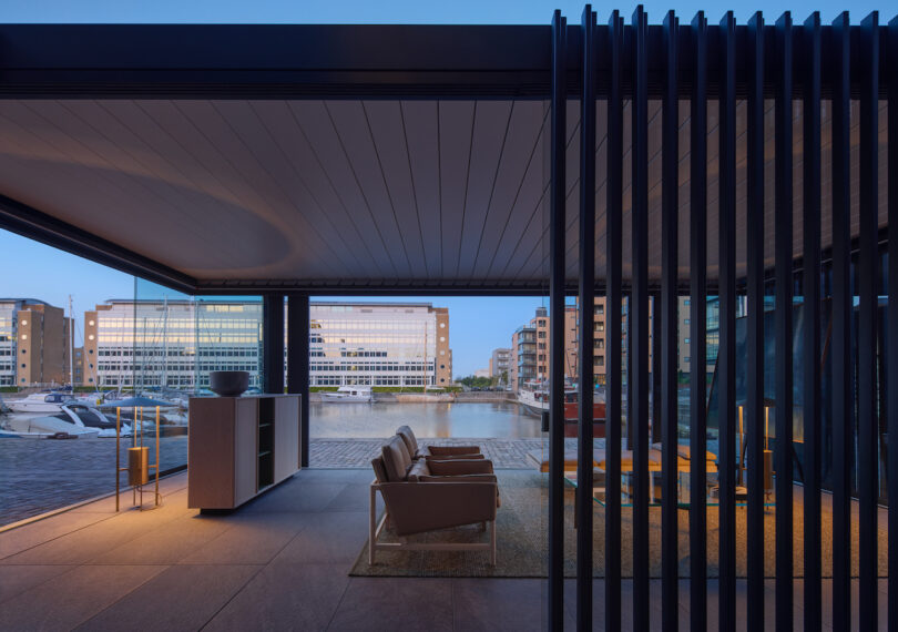 Modern open-air lounge area with two armchairs overlooks a waterfront marina and office buildings at dusk, separated by vertical black slats