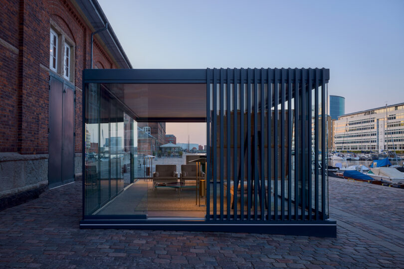 A modern glass-walled pavilion with vertical metal slats sits on a cobblestone waterfront, surrounded by industrial and office buildings