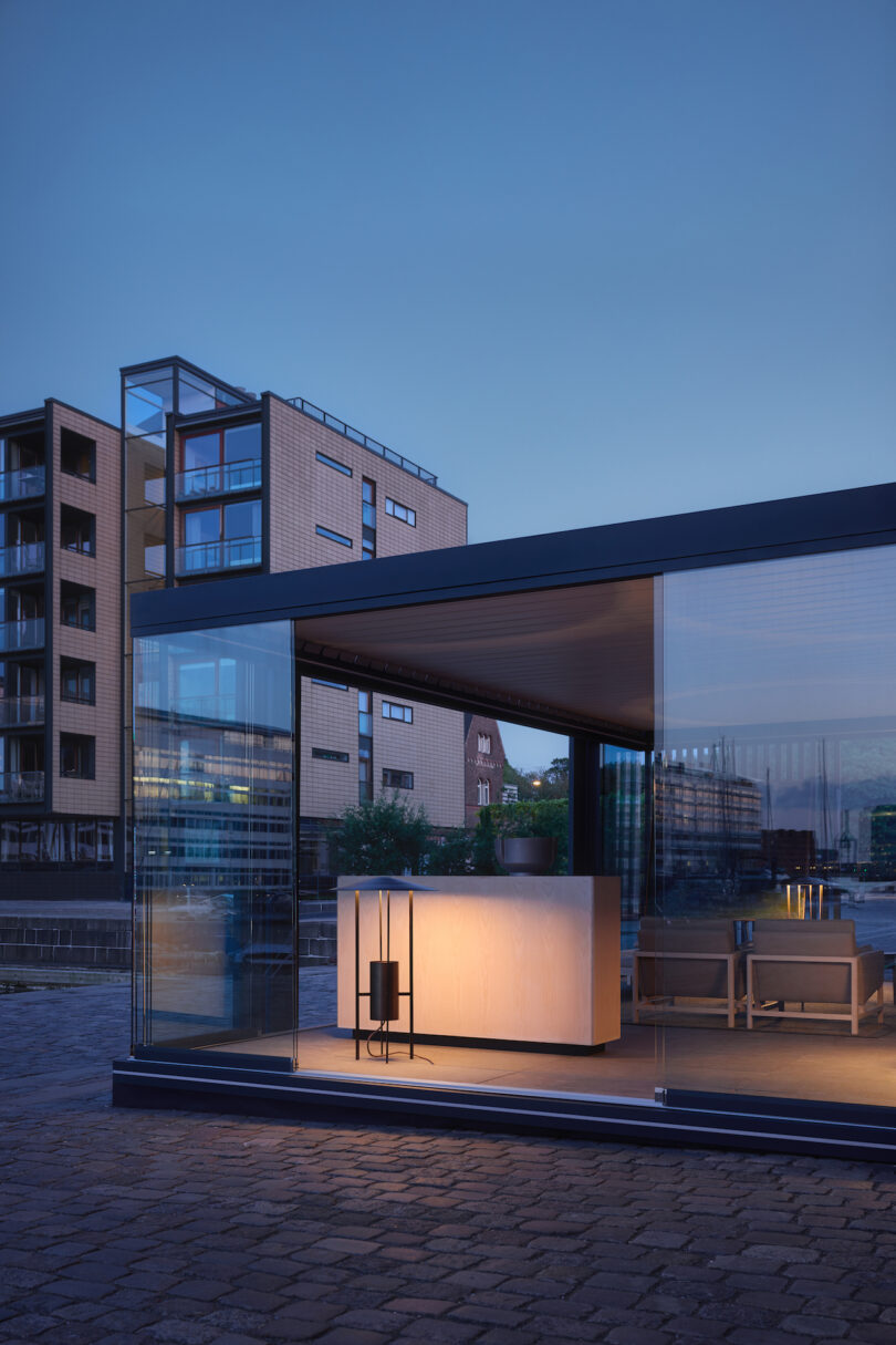 Modern glass-walled building with interior lights on at dusk, adjacent to brick apartments and a waterfront area