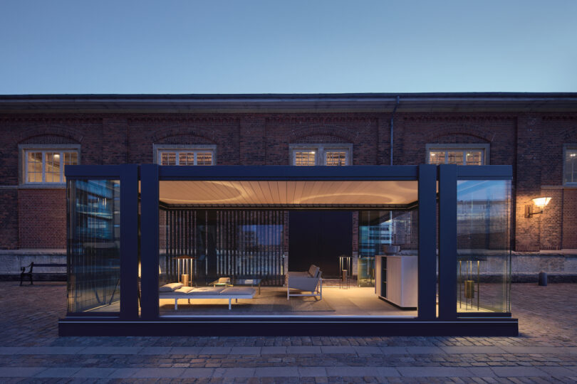 A modern glass-walled room with minimalist furniture is illuminated at dusk, set in front of a historic brick building with arched windows