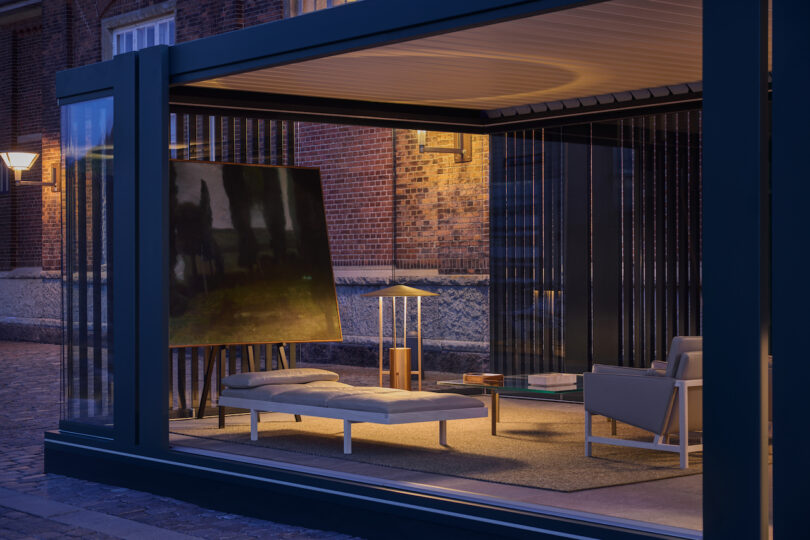Modern glass-walled room at dusk with a daybed, armchair, standing lamp, side tables, and a large abstract painting on an easel against a backdrop of brick buildings