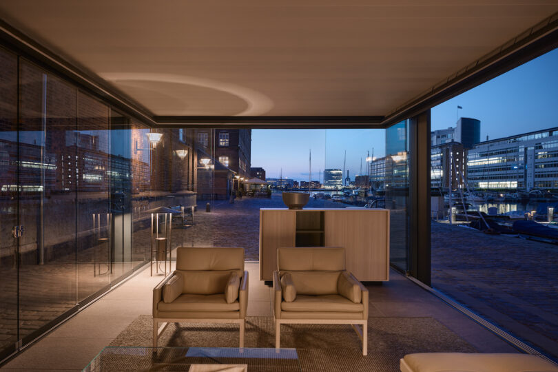 Modern indoor lounge with two beige armchairs and a sideboard, featuring large glass windows overlooking a harbor and cityscape at dusk