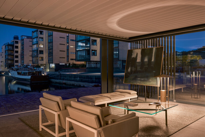 Modern living room with beige furniture and large windows overlooking a canal with a docked boat and contemporary apartment buildings at dusk