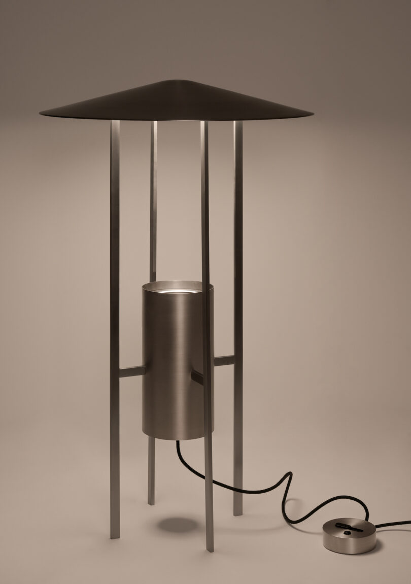 A modern metal floor lamp with a wide, cone-shaped shade, cylindrical base, four vertical supports, and a cord connected to a round power switch