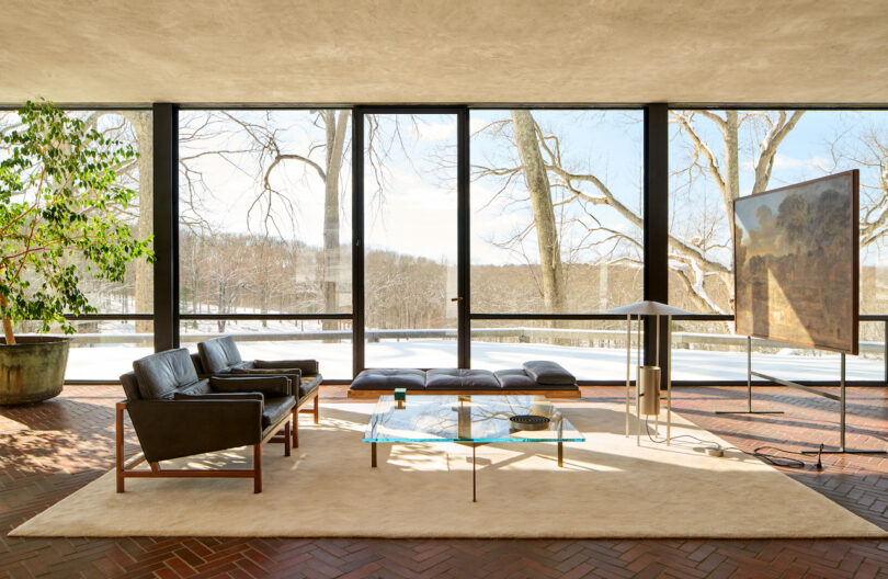 A modern living room with glass walls, black leather chairs, a glass coffee table, large painting, and a view of snowy trees outside