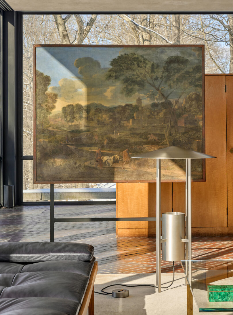 A large landscape painting is displayed on a stand in a modern living room with floor-to-ceiling windows, wood paneling, and a metallic floor lamp in the foreground