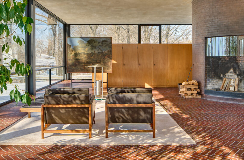 Two modern armchairs face a large painting in a bright room with floor-to-ceiling windows, brick flooring, a wood-burning fireplace, and stacked firewood