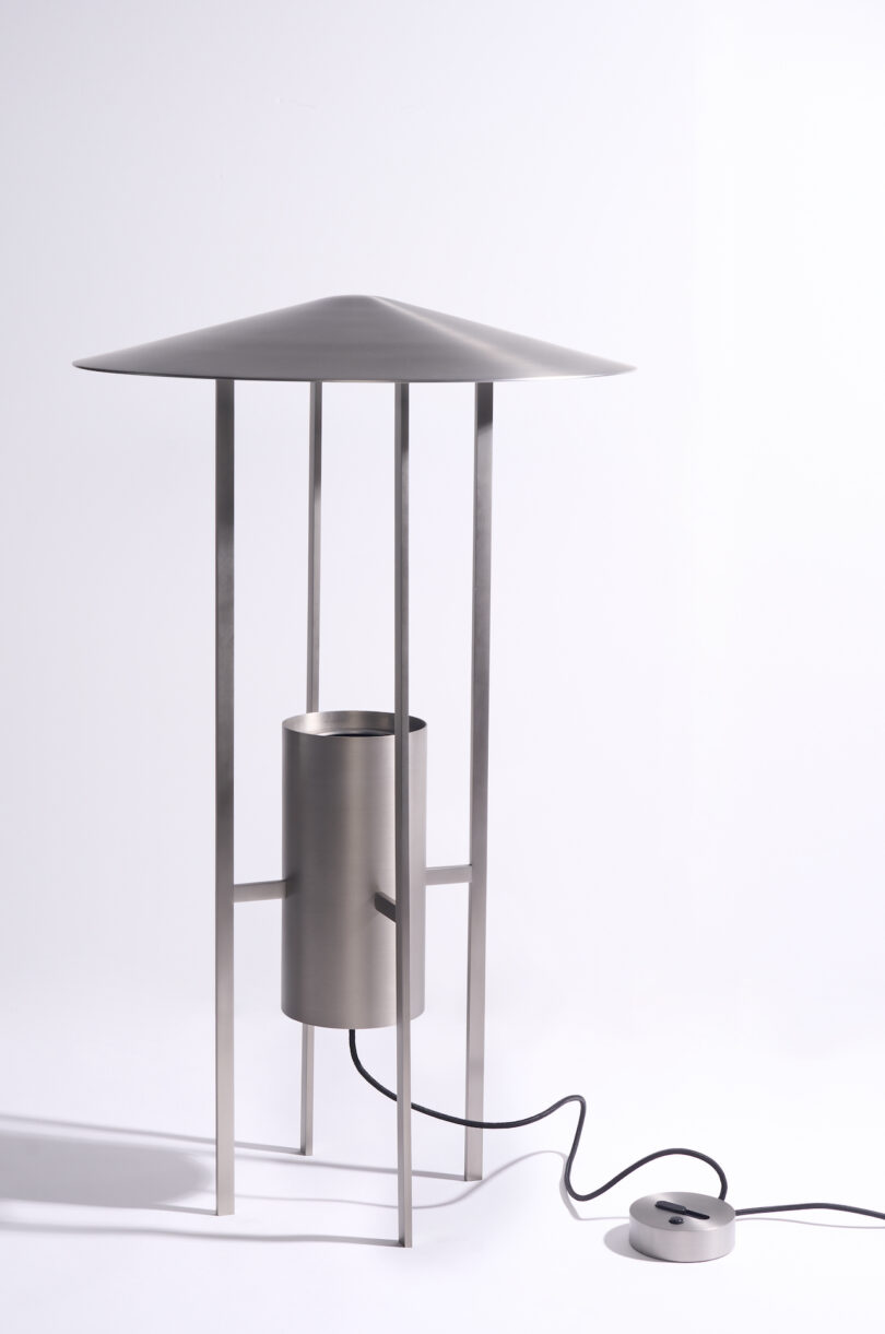 A modern metallic lamp with a wide, conical shade, four vertical legs, a cylindrical base, and a visible power cord connected to a round switch