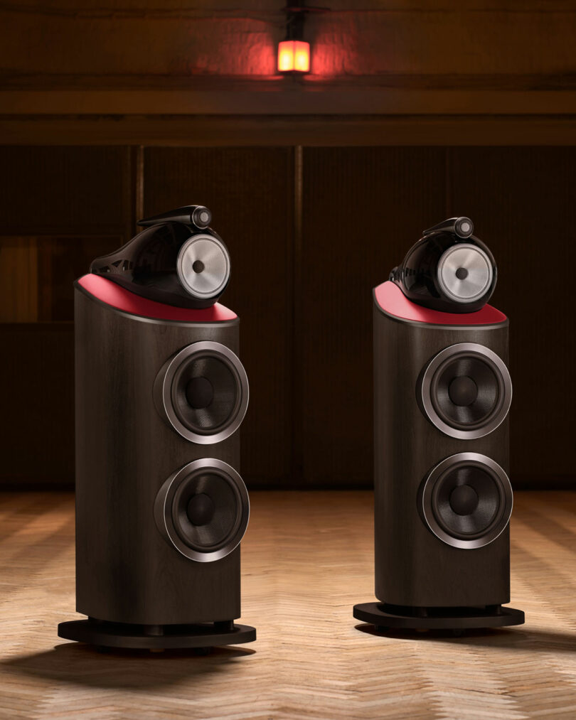 Two high-end Bowers & Wilkins speakers with a dark wood finish and red accents are positioned on a wooden floor in a dimly lit room.
