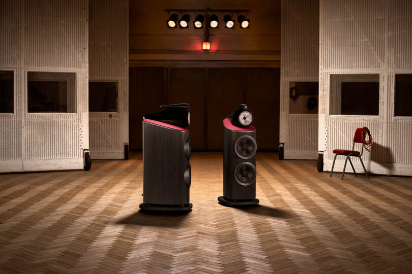 Two large, high-end Bowers & Wilkins speakers are placed on a herringbone wood floor in a dimly lit room with soundproofed walls, a red chair, and overhead stage lights.