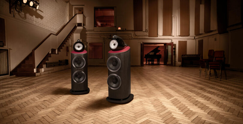 Two large Bowers & Wilkins black speakers are positioned in the center of a spacious recording studio with parquet flooring, a grand piano in the back, and stacked chairs against the wall.
