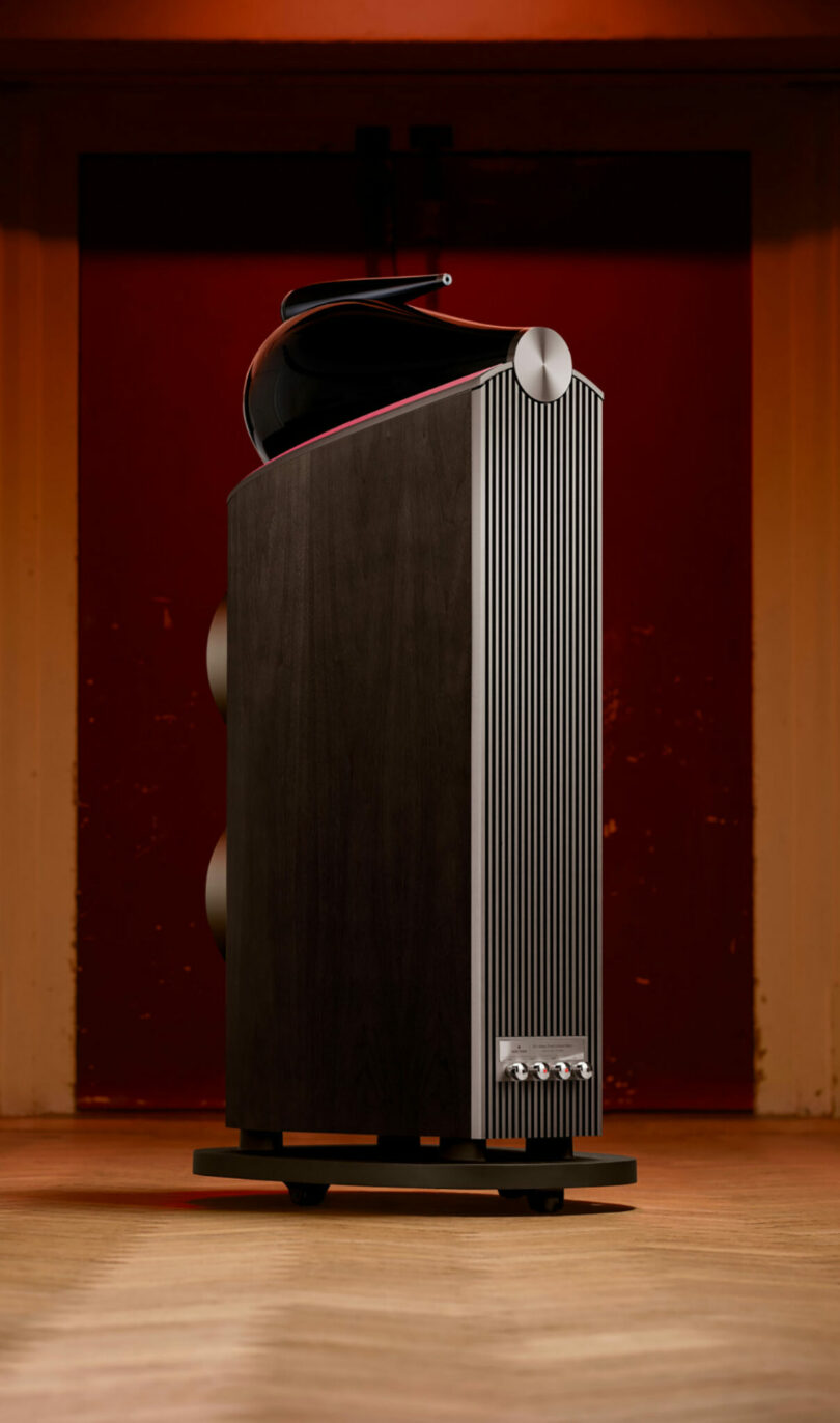 Tall, modern Bowers & Wilkins floor-standing speaker with a dark wood finish, vertical metallic accents, and a curved top element, set against a red and orange background.