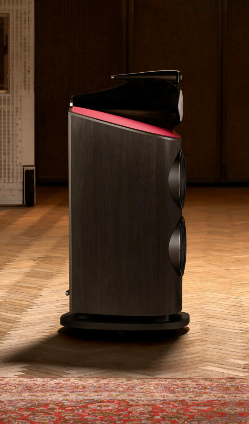 A large, modern black Bowers & Wilkins speaker with a red accent is positioned on a wooden floor next to a patterned rug, against a background of acoustic panels.
