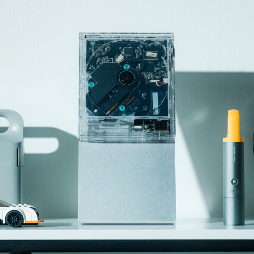 La electrónica transparente con componentes internos visibles se encuentran en un estante al lado de un pequeño automóvil de juguete, con objetos cilíndricos grises y amarillos.