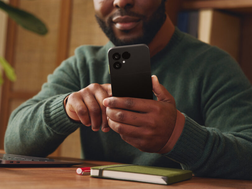 A person in a green sweater sits at a desk holding a black Fairphone, with a closed notebook and pen in front of them.