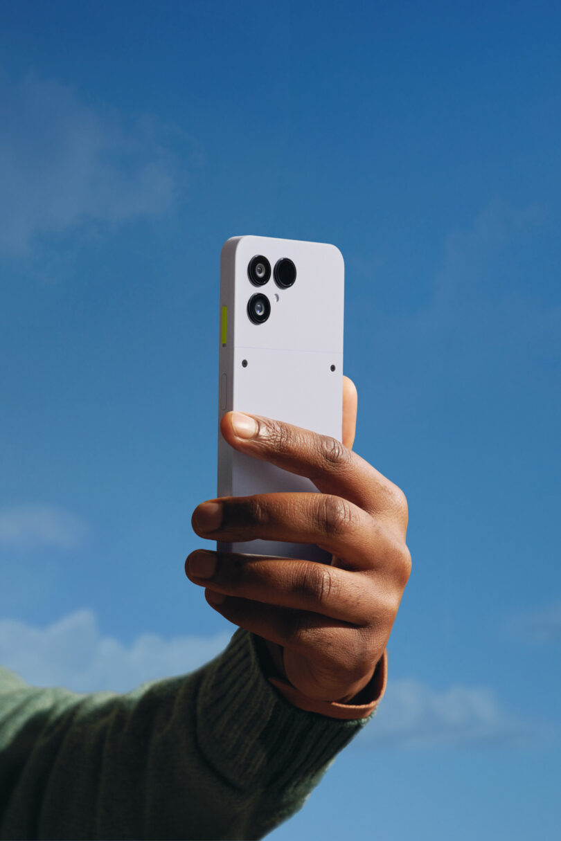 A person holds up a white Fairphone smartphone with three rear cameras against a clear blue sky.