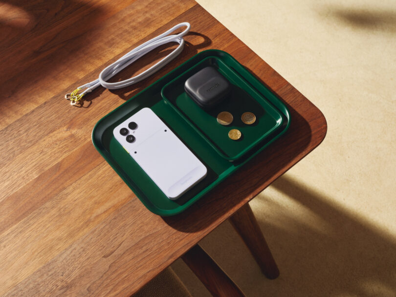 A white Fairphone, a pair of black wireless earbuds in a case, and three coins sit in a green tray on a wooden table next to a white charging cable.