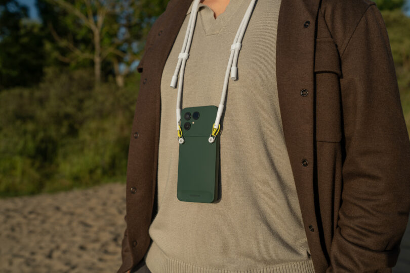 Person outdoors wearing a brown sweater and beige shirt with a green Fairphone attached to a white lanyard hanging from their neck. Trees and sand are visible in the background.