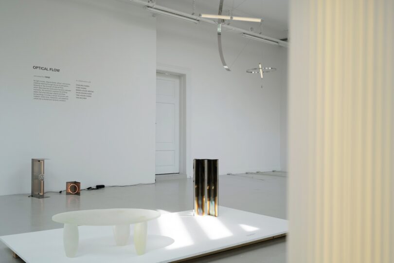 A minimal gallery space with modern abstract sculptures, including a white oval table, cylindrical objects, and metallic hanging pieces, titled "Optical Flow" on the wall.