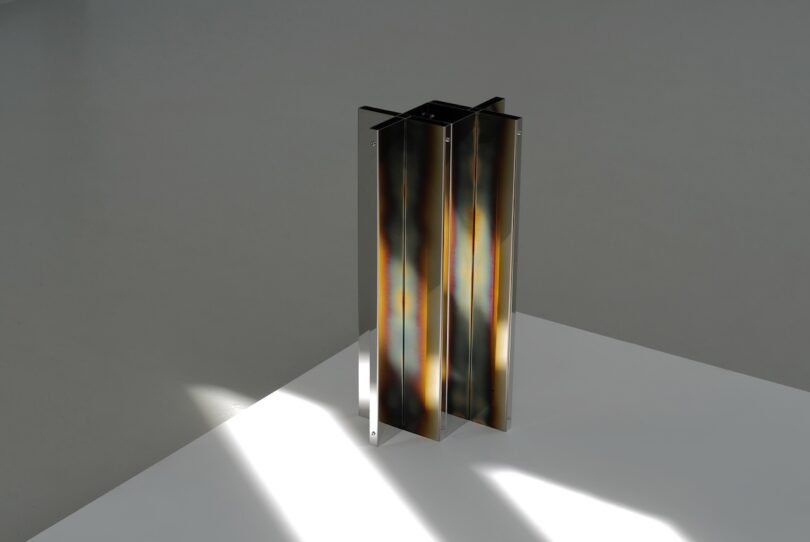 A metallic geometric sculpture with vertical, reflective panels stands on a white and gray surface, illuminated by natural light.
