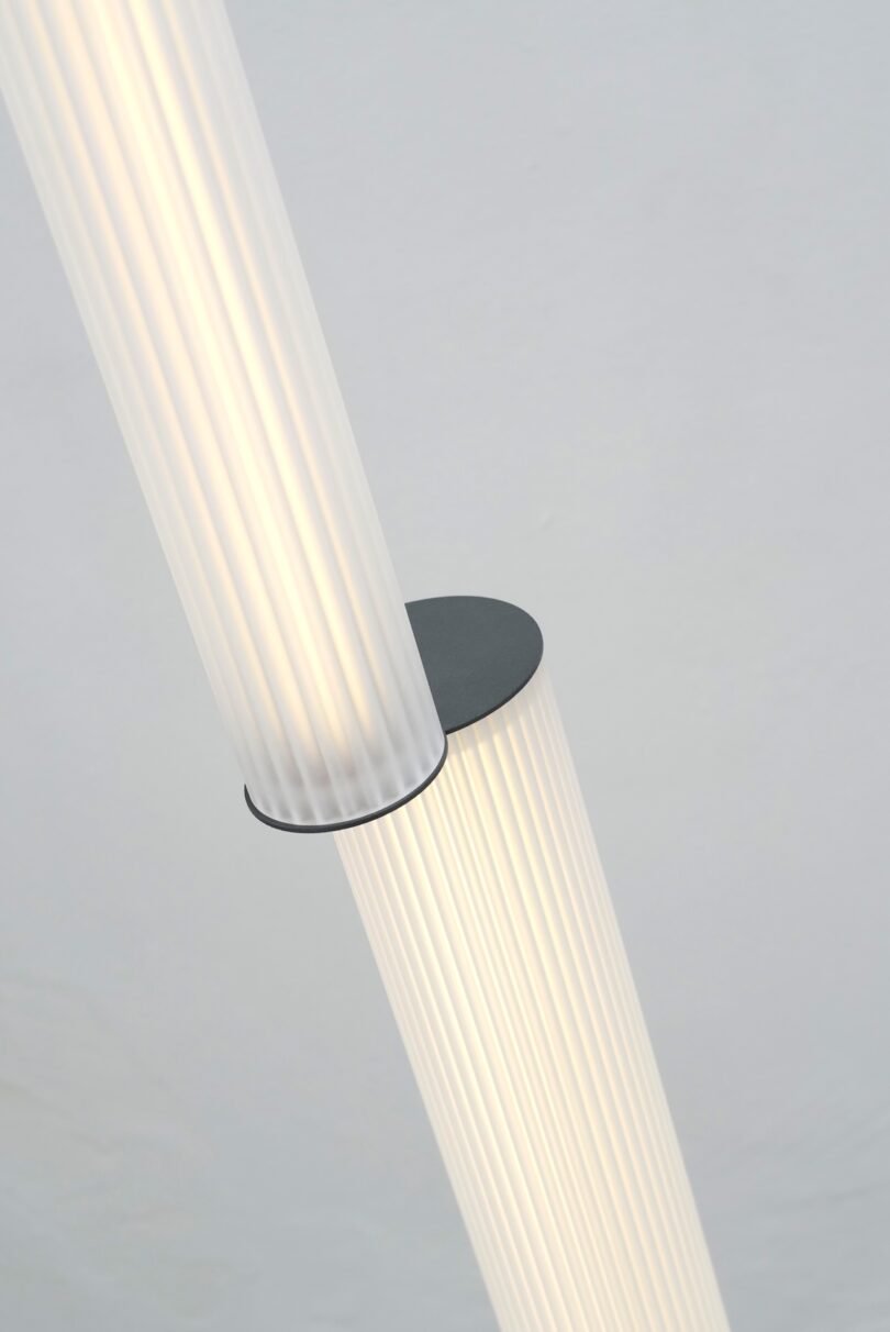 A close-up of two intersecting, vertically ribbed, FANZI cylindrical light fixtures with a round, black connecting element against a plain light background.