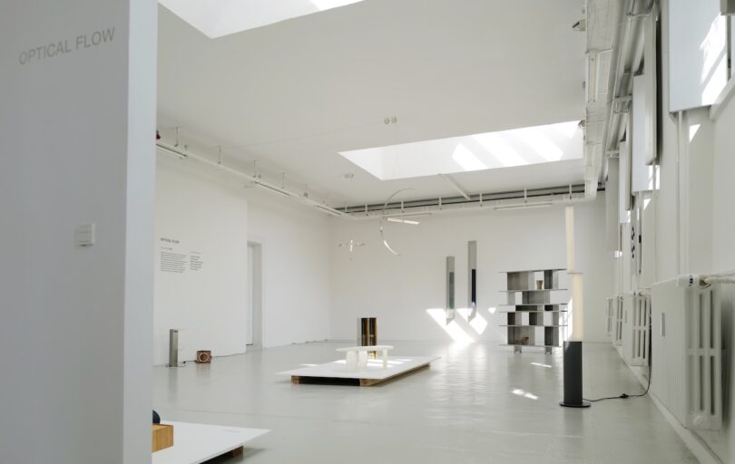 Minimalist contemporary art exhibition in a white gallery space with sculptures and installations displayed on the floor and natural light streaming through skylights.