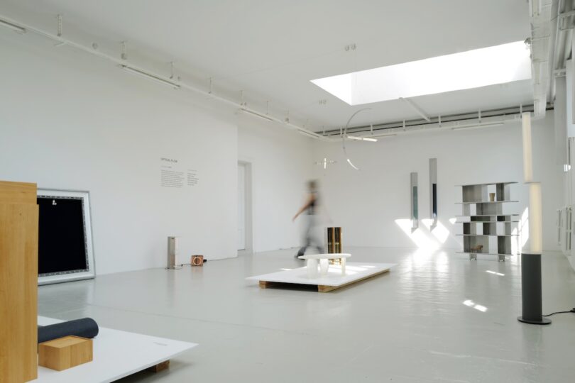 A minimal, white-walled gallery space with modern art installations and a blurred figure walking in the background.