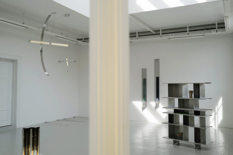 A modern, minimalist gallery space with geometric light fixtures, metallic shelving, and tall vertical mirrors, divided by a central column.