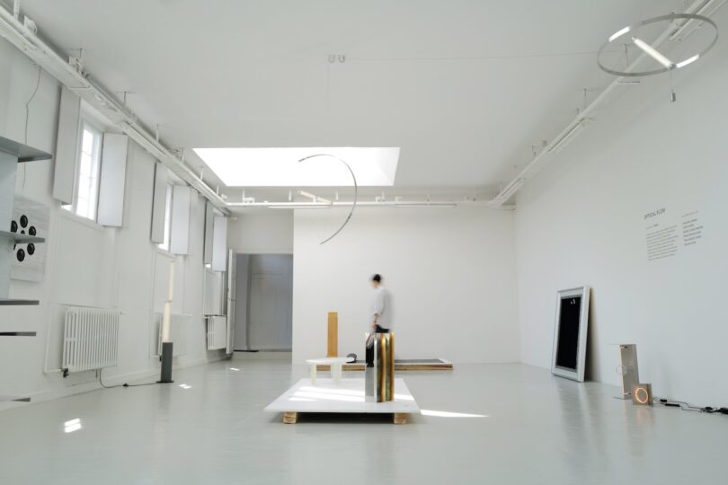 A person stands in a bright, minimalist art gallery featuring abstract sculptures and wall installations on a white floor.