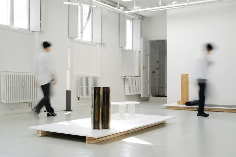 A minimal art gallery with abstract sculptures on white platforms and two blurred figures walking through the bright, white space.