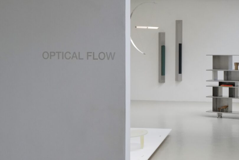 A minimal art gallery interior with the words "OPTICAL FLOW" on a gray wall; shelves and wall-mounted pieces are visible in the background.