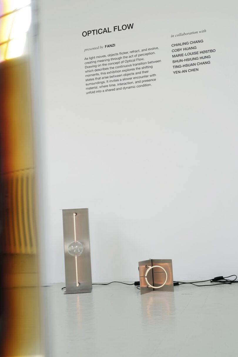 A gallery installation features two minimalist metal and glass FANZI light sculptures on the floor near a white wall with text describing the exhibit "Optical Flow.