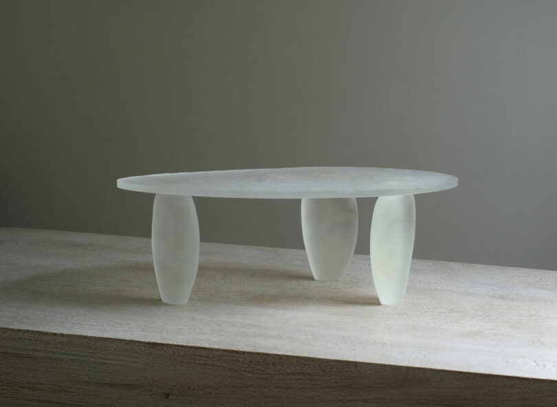 A translucent, oval-shaped FANZI table with three rounded, frosted glass legs sits on a textured, light-colored surface against a plain gray background.