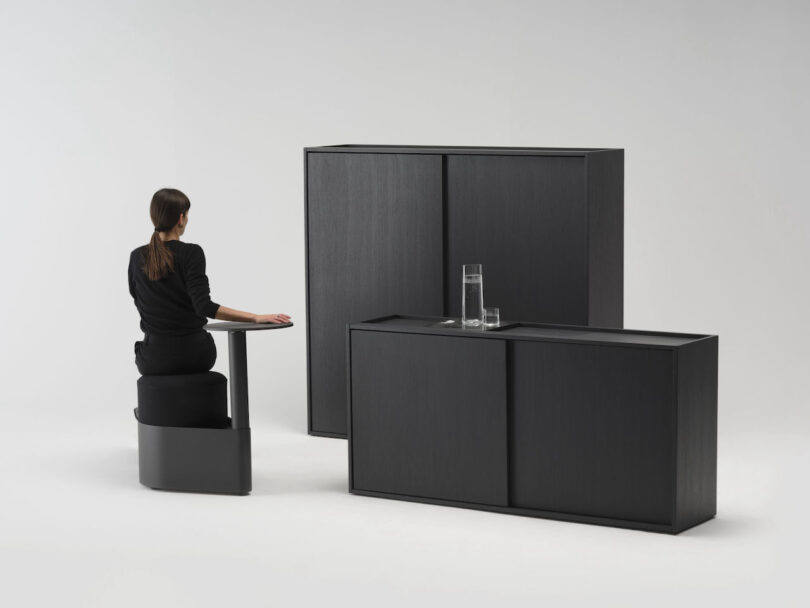 A woman in black sits at a minimalist black desk with matching cabinets, one holding a glass and a carafe of water, against a plain white background