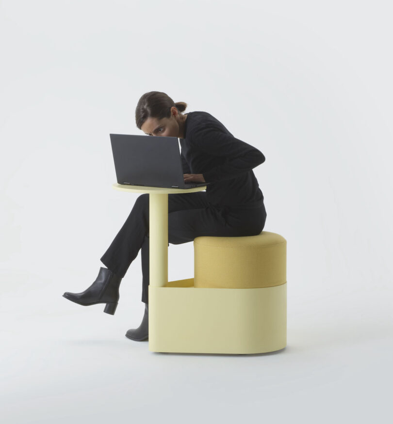 A person in black attire sits on a round yellow stool, working on a laptop at a matching yellow desk, against a plain white background