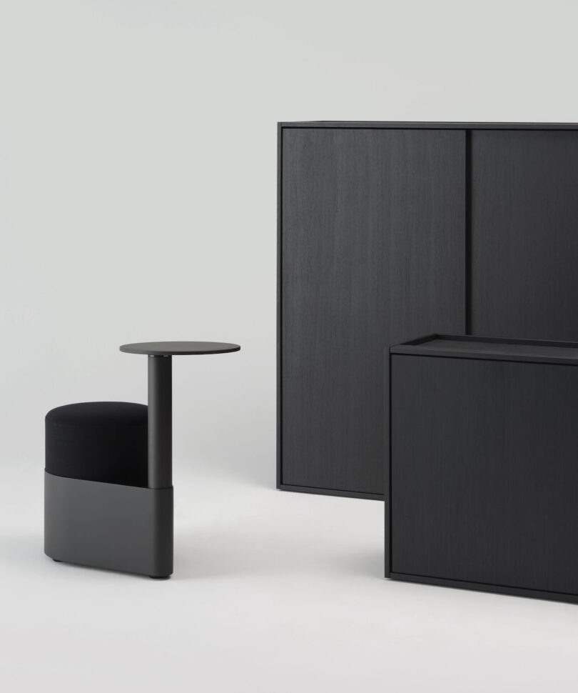 A black, minimalist chair with an attached round side table sits next to two black rectangular cabinets against a plain light gray background
