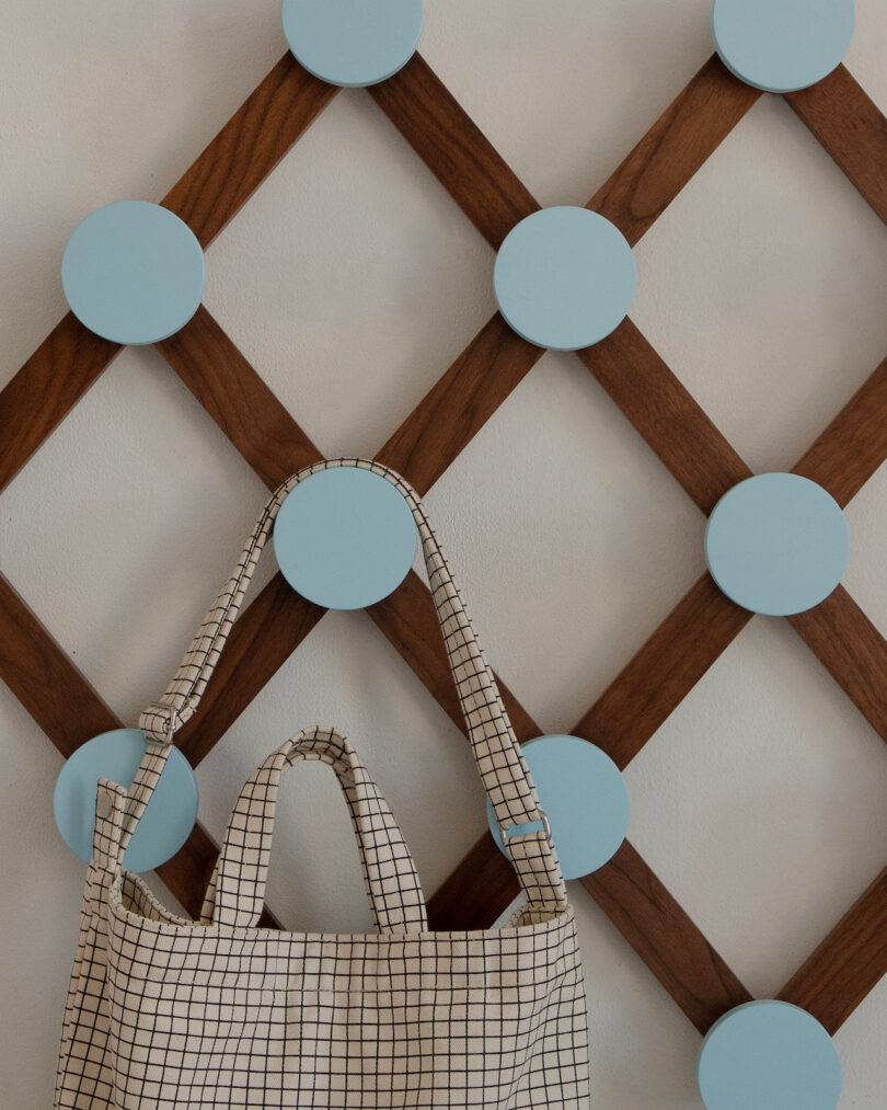 A beige checkered tote bag hangs on a wooden wall rack with intersecting slats and large, round, light blue pegs