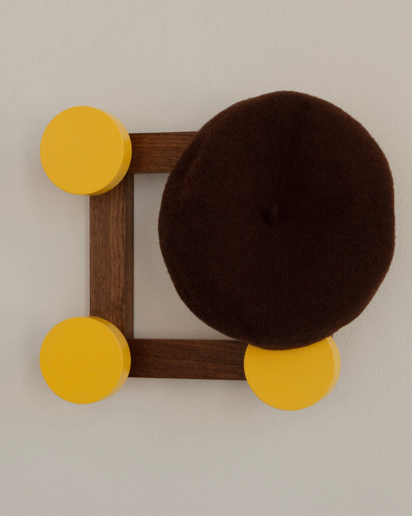 A brown beret hangs on a wall-mounted wooden square with four round yellow hooks at each corner