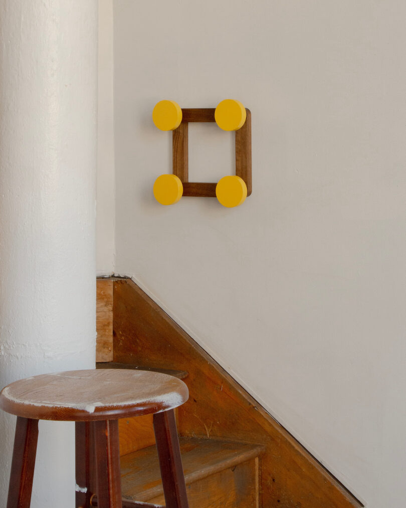 A wooden stool sits by a staircase with a wall-mounted, square wooden decor featuring four yellow circular ends