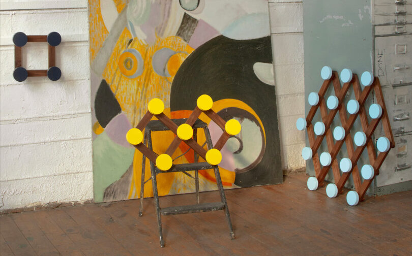 Geometric wall sculptures with circular nodes by Hanna Anonen are displayed in a studio space with abstract paintings, a chair, and a metal cabinet on a wooden floor.