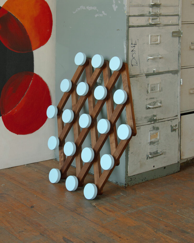 A wooden lattice structure with light blue circular pads leans against a metal filing cabinet in a room with wooden floors and abstract wall art