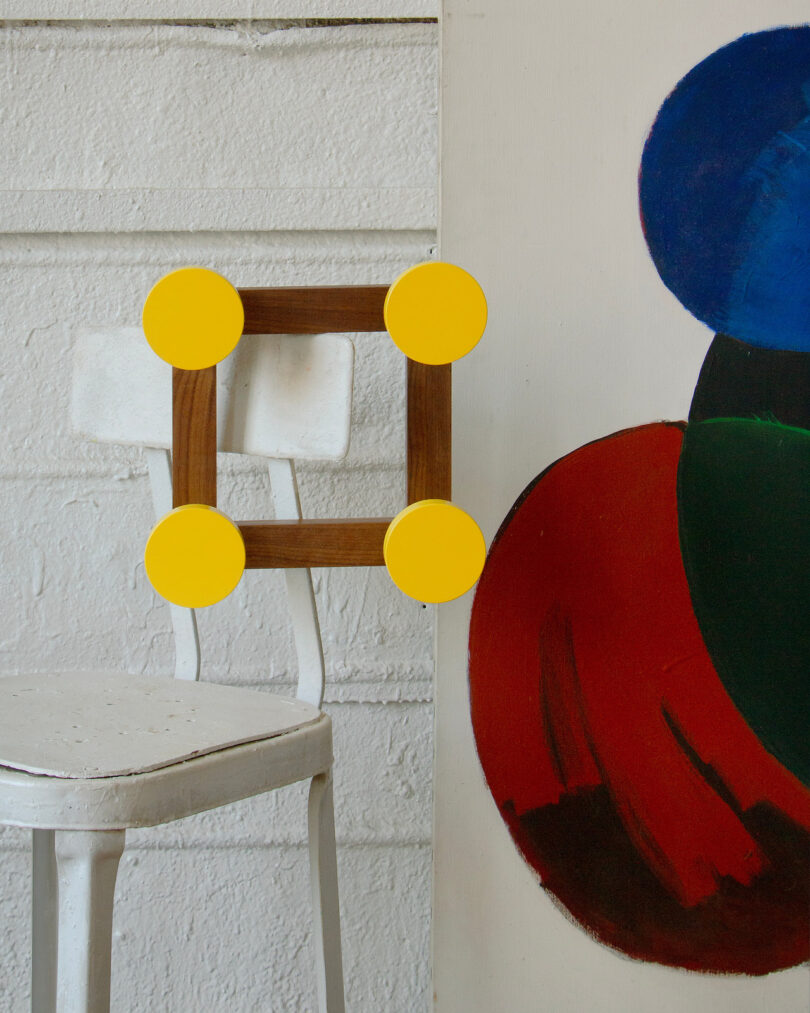 A white metal chair is next to a colorful abstract painting. A wooden square with yellow circles at each corner rests on the chair's back