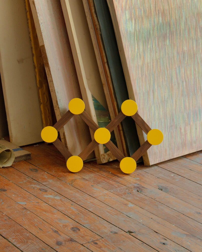 A geometric wooden structure with yellow circular ends leans against stacked canvases on a wooden floor