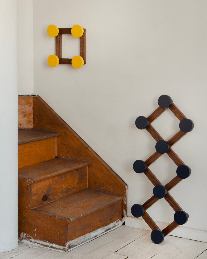 A small set of wooden stairs beside two geometric wall sculptures; one is square with yellow circles and one is diamond-shaped with dark circles