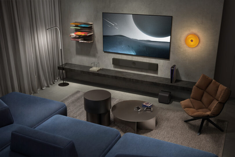 Modern living room with blue sectional sofa, brown swivel chair, KEF XIO speakers, round coffee tables, wall-mounted TV displaying a sci-fi scene, and minimalist decor.